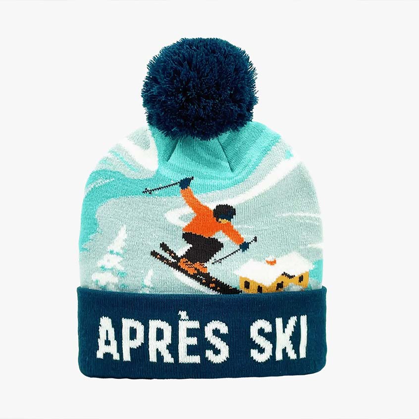 A knit pom-pom beanie with a dark navy blue top and cuff, featuring a light blue and white ski-themed graphic with a skier and the text 'APRÈS SKI' in white on the cuff.