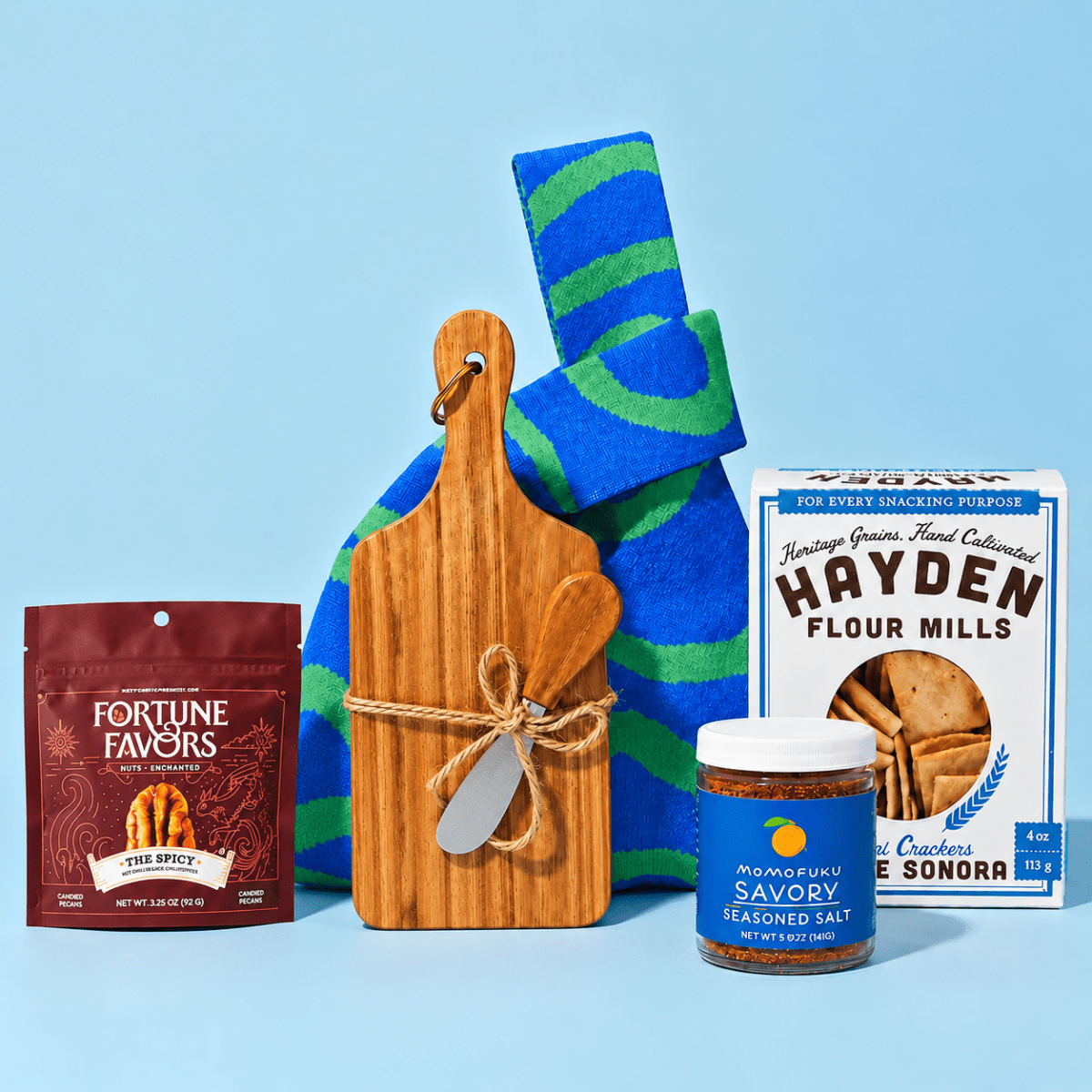 Gourmet gift collection featuring Fortune Favors spicy pecans, a bamboo cutting board with cheese knives, a blue and green patterned towel, Hayden Flour Mills crackers, and Momofuku savory seasoned salt, arranged against a light blue background.