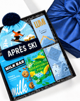 A curated gift box set featuring a light blue knit beanie with a dark blue pom-pom and ski graphic, a blue après ski tag, a chocolate chip milk bar, sour elderflower fish crackers, and a blue satin gift bag on a white background.