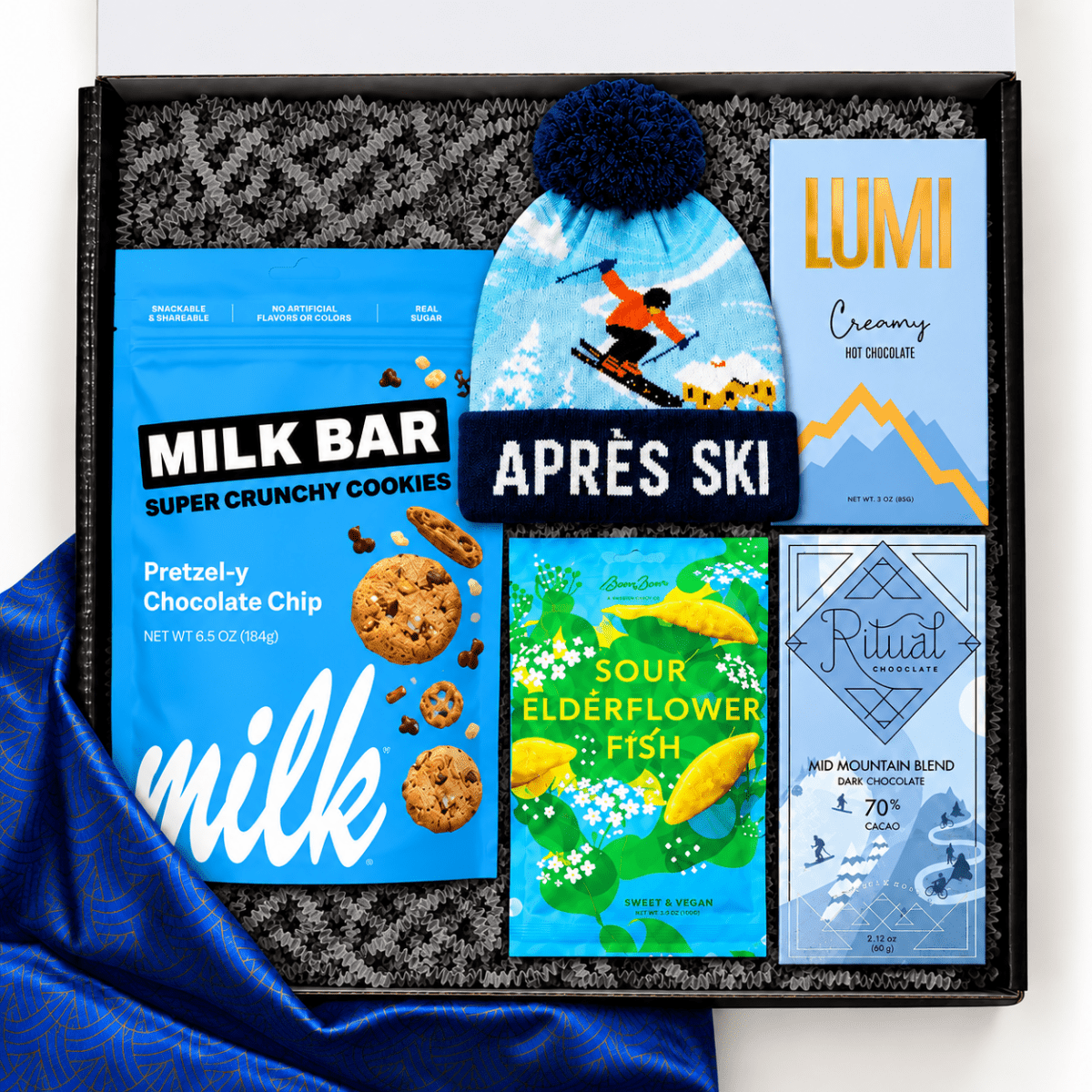 Après-ski gift box featuring a blue ski-themed pom-pom beanie, Milk Bar Pretzel-y Chocolate Chip cookies, Lumi creamy hot chocolate, BonBon Sour Elderflower Fish candy, and Ritual dark chocolate, arranged in a black gift box with gray crinkle paper and blue patterned fabric.