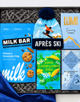Après-ski gift box featuring a blue ski-themed pom-pom beanie, Milk Bar Pretzel-y Chocolate Chip cookies, Lumi creamy hot chocolate, BonBon Sour Elderflower Fish candy, and Ritual dark chocolate, arranged in a black gift box with gray crinkle paper and blue patterned fabric.