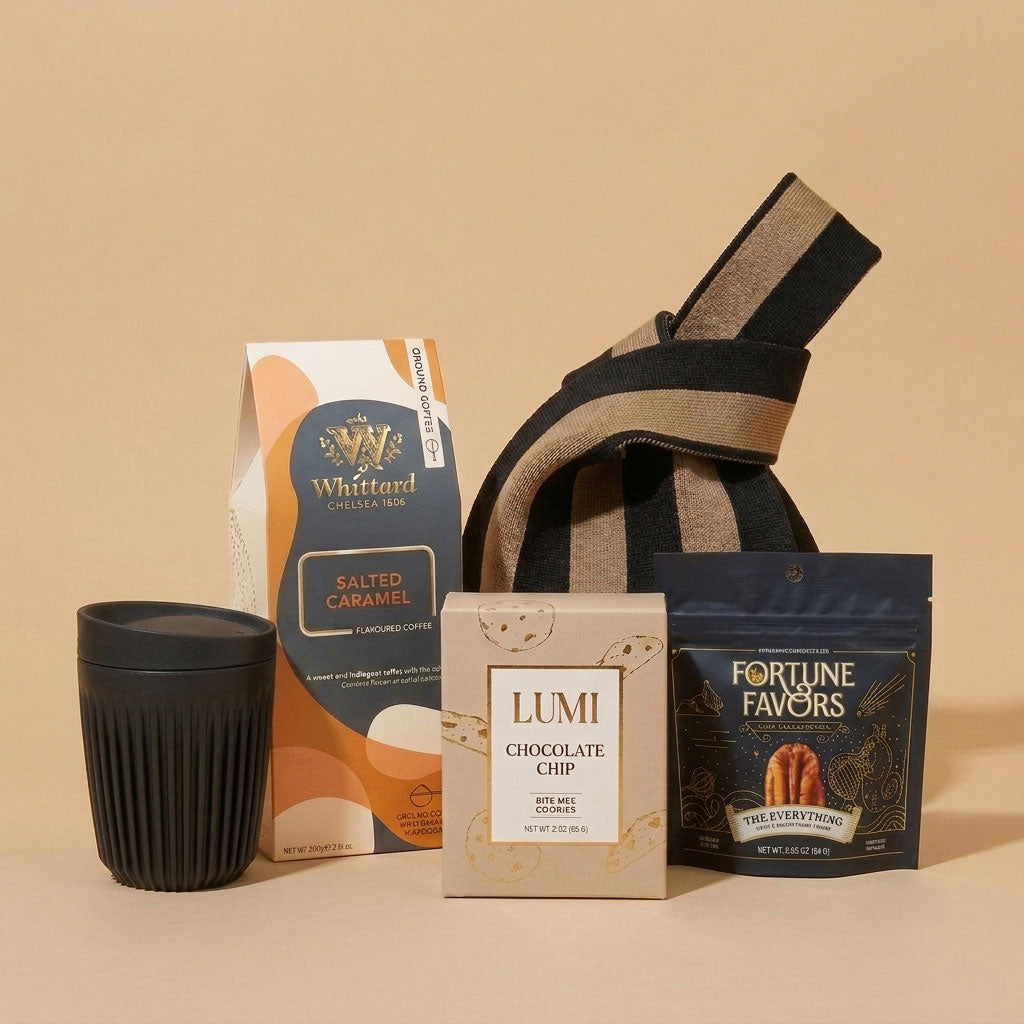 A curated gift set on a warm beige background, featuring a black travel tumbler, a Whittard Salted Caramel ground coffee bag, a box of Lumi Chocolate Chip cookies, a Fortune Favors 'One Everything' bag, and a striped fabric tote bag.