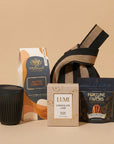 A curated gift set on a warm beige background, featuring a black travel tumbler, a Whittard Salted Caramel ground coffee bag, a box of Lumi Chocolate Chip cookies, a Fortune Favors 'One Everything' bag, and a striped fabric tote bag.