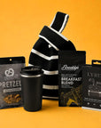 A curated gift bag set featuring a black and white striped knit bag, a black travel tumbler, a bag of Brooklyn Roastery Breakfast Coffee, a bag of On Tap Kitchen Pretzels, and a boxed pair of LYRIX wireless earbuds, arranged on a solid mustard yellow background.