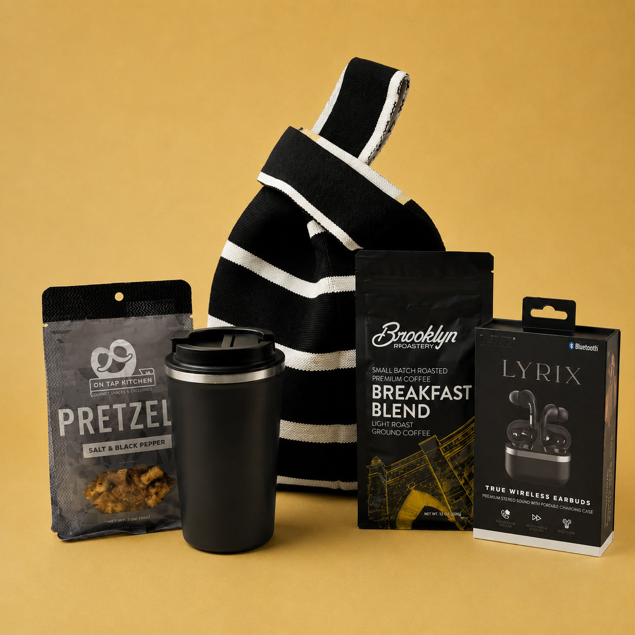 A curated gift bag set featuring a black and white striped knit bag, a black travel tumbler, a bag of Brooklyn Roastery Breakfast Blend Coffee, a bag of On Tap Kitchen Pretzels, and a boxed pair of LYRIX wireless earbuds, arranged on a solid mustard yellow background.