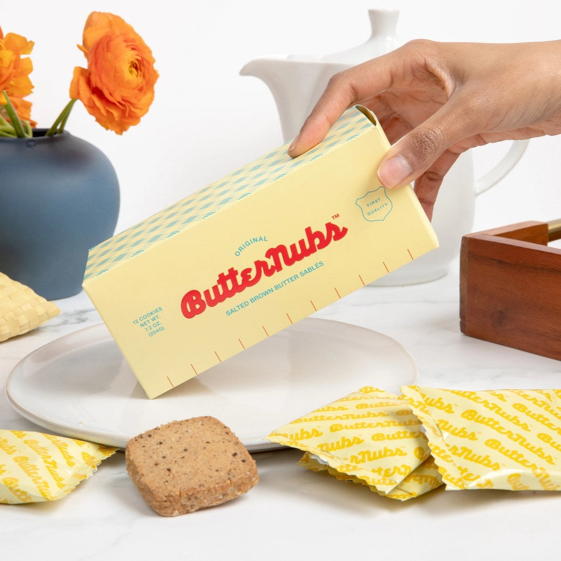 A hand holds a Butternubs butter package over a white plate, with several yellow-and-white ButterTubs wrapper pieces and a brown cookie on a white tabletop. A blue vase with orange flowers and a white teapot are blurred in the background.