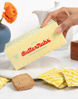 A hand holds a Butternubs butter package over a white plate, with several yellow-and-white ButterTubs wrapper pieces and a brown cookie on a white tabletop. A blue vase with orange flowers and a white teapot are blurred in the background.