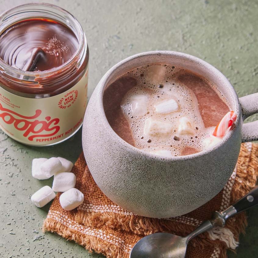 A ceramic mug filled with hot chocolate topped with marshmallows and a candy cane, next to an open jar of Coops's Peppermint Hot Fudge on a textured surface.