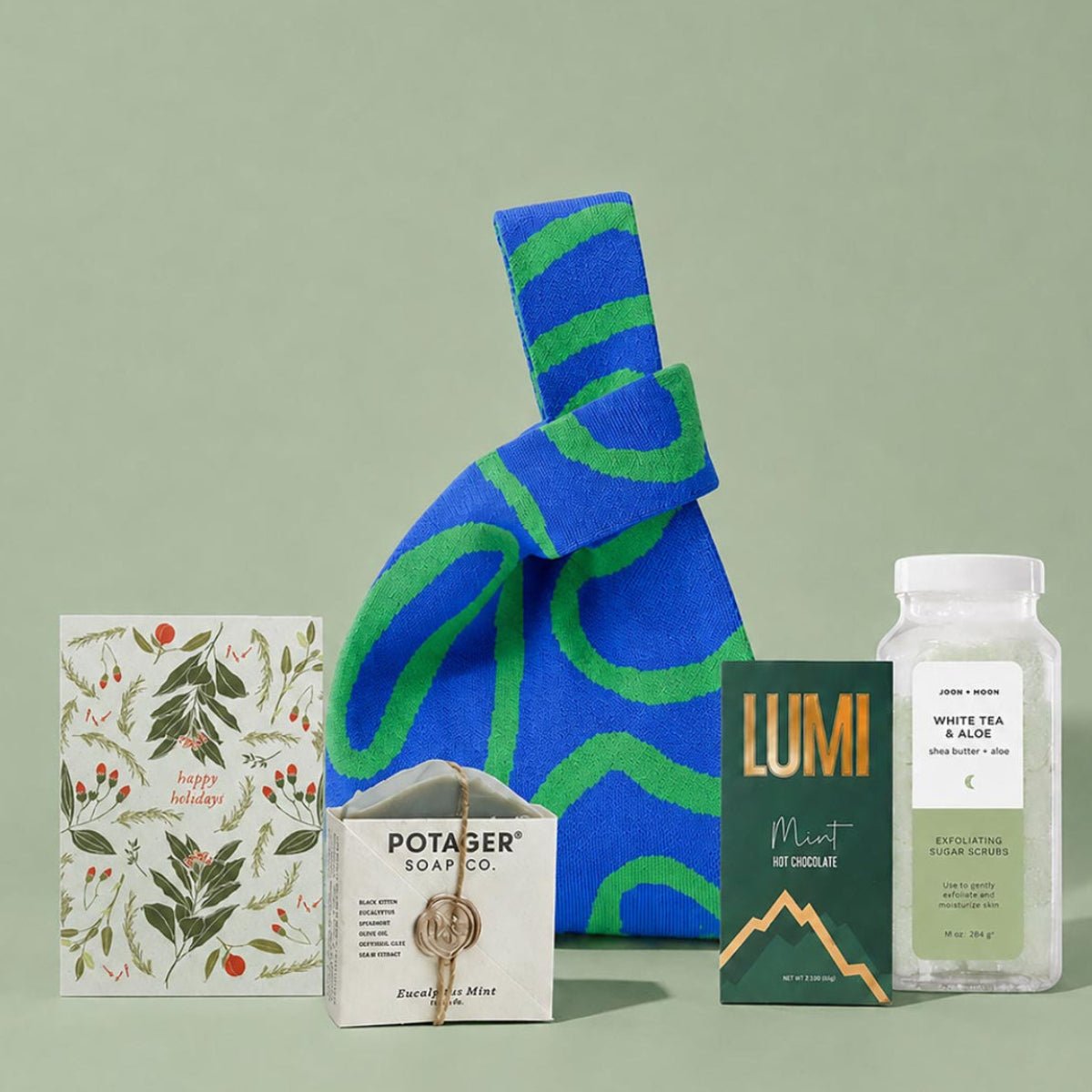 A curated gift bag set arranged on a pale green background, featuring a large blue and green patterned knit bag, a holiday card, a bar of Potager body co. soap, a box of Lumi Mint hot chocolate mix, and a clear bottle of Joon Moon White Tea & Aloe Sugar Scrub