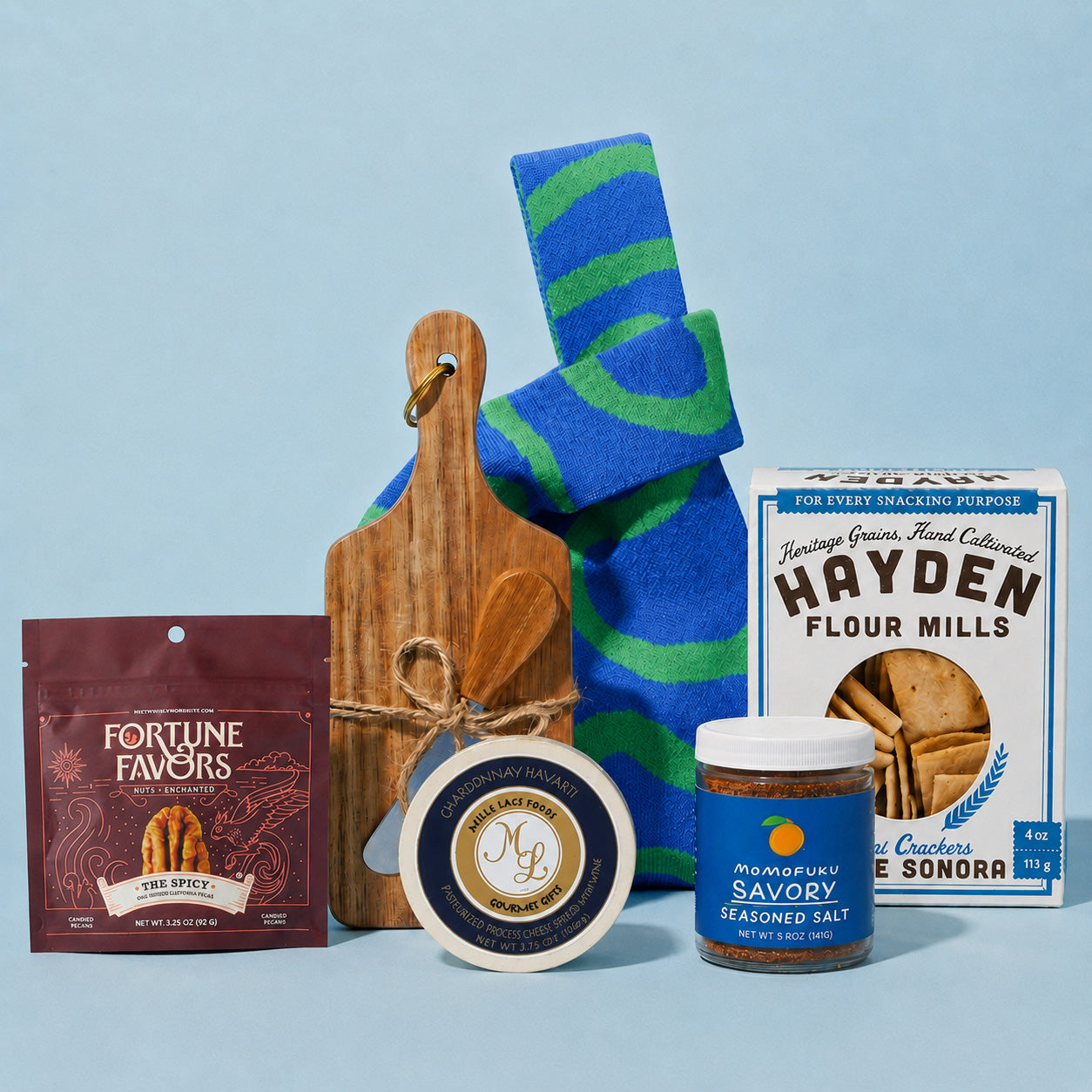 A curated snack and food gift set arranged on a light blue background, including a package of Fortune Favors candied pecans, a round cheese wheel, a jar of Momofuku savory seasoned salt, a box of Hayden Flour Mills crackers, and a wooden cutting board with a striped blue and green swirl patterned knit bag behind it.