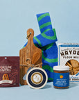 A curated snack and food gift set arranged on a light blue background, including a package of Fortune Favors candied pecans, a round cheese wheel, a jar of Momofuku savory seasoned salt, a box of Hayden Flour Mills crackers, and a wooden cutting board with a striped blue and green swirl patterned knit bag behind it.