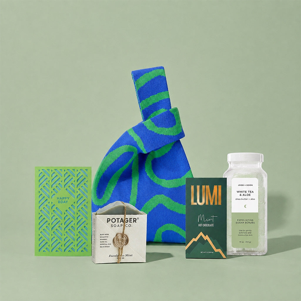 A curated gift bag set arranged on a pale green background, featuring a large blue and green patterned knit bag, a green cut out birthday card, a bar of Potager body co. soap, a box of Lumi Mint hot chocolate mix, and a clear bottle of Joon Moon White Tea & Aloe Sugar Scrub