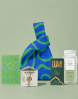 A curated gift bag set arranged on a pale green background, featuring a large blue and green patterned knit bag, a green cut out birthday card, a bar of Potager body co. soap, a box of Lumi Mint hot chocolate mix, and a clear bottle of Joon Moon White Tea & Aloe Sugar Scrub