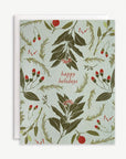 A rectangular holiday greeting card with a botanical pattern of green leaves and red berries on a pale blue-green background, with the words 'happy holidays' in red script. A white envelope is partially visible behind it on a light neutral background.