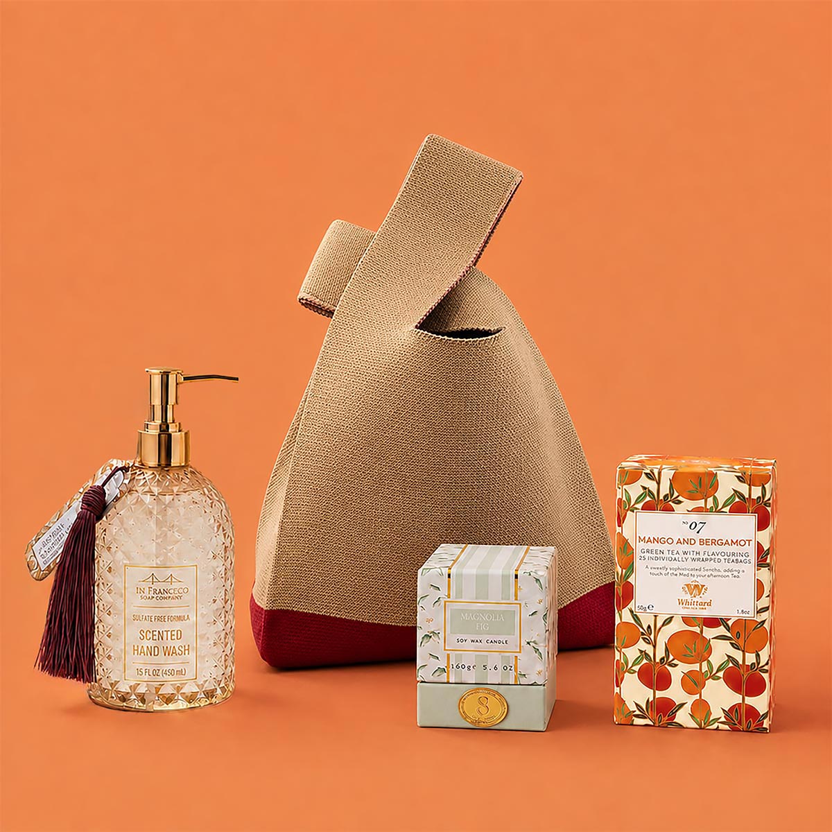 A curated gift bag set featuring a beige knit bag with a dark red base, a glass bottle of Jasmine and Sandalwood Hand Soap with gold hardware and a tassel, a small box of soy wax magnolia fig candle, and a box of Whittard Mango and Bergamot tea, all arranged on a solid orange background. 