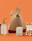 A curated gift bag set featuring a beige knit bag with a dark red base, a glass bottle of Jasmine and Sandalwood Hand Soap with gold hardware and a tassel, a small box of soy wax magnolia fig candle, and a box of Whittard Mango and Bergamot tea, all arranged on a solid orange background. 