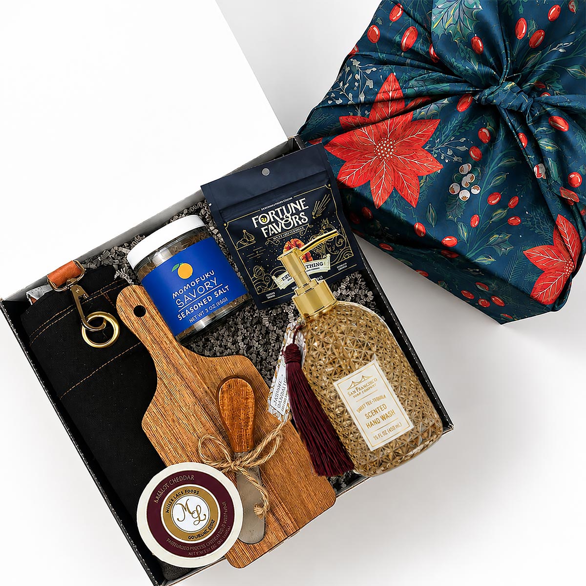 A curated gift box set shown from above on a white background, including a black apron with gold lettering, a blue jar of Momofuku seasoned salt, a packet of Fortune Favors candied pecans, a bottle of hand soap with a gold pump and tassel, a wooden cutting board and cheese spread knife tied with twine, and a wheel of cheese. A green gift wrap with red floral patterns is partially visible at the top right.

