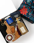 A curated gift box set shown from above on a white background, including a black apron with gold lettering, a blue jar of Momofuku seasoned salt, a packet of Fortune Favors candied pecans, a bottle of hand soap with a gold pump and tassel, a wooden cutting board and cheese spread knife tied with twine, and a wheel of cheese. A green gift wrap with red floral patterns is partially visible at the top right.

