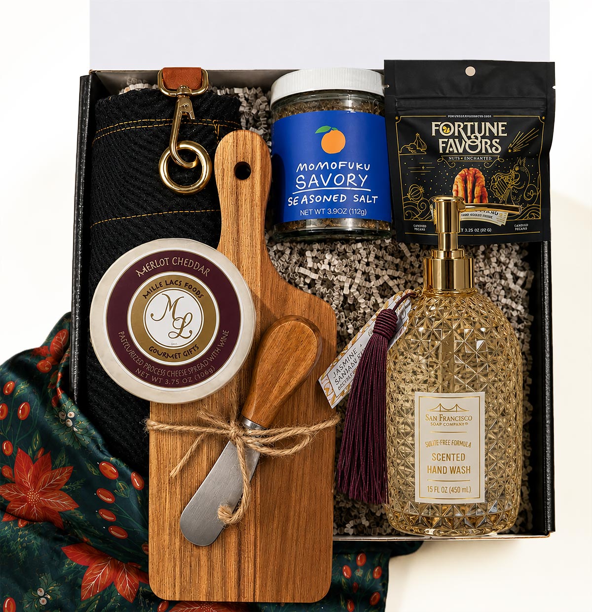 A curated gift box set shown from above containing a wooden cutting board and cheese spread knife tied with twine, a bottle of scented hand wash with a gold pump and tassel, a jar of seasoned salt, a bag of Fortune Favors candied pecans, a wheel of cheese and a dark apron, arranged in a box.