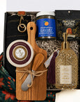 A curated gift box set shown from above containing a wooden cutting board and cheese spread knife tied with twine, a bottle of scented hand wash with a gold pump and tassel, a jar of seasoned salt, a bag of Fortune Favors candied pecans, a wheel of cheese and a dark apron, arranged in a box.