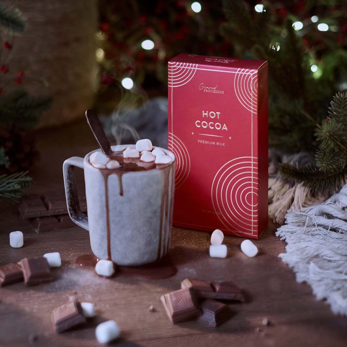 Hot chocolate with marshmallowss, a box of Grand Provisions hot cocoa, and chocolate pieces on a wooden surface with a Christmas tree in the background.