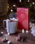 Hot chocolate with marshmallowss, a box of Grand Provisions hot cocoa, and chocolate pieces on a wooden surface with a Christmas tree in the background.