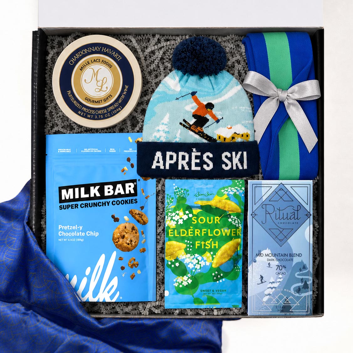 A curated gift box set labeled 'APRÈS SKI' containing a light blue knit beanie with a dark blue pom-pom, a blue and green striped knitted mini bag tied with a silver ribbon, a tin of Cheesecake &amp; Honey spreads, a bag of Milk Bar pretzel chocolate chip cookies, a bag of sour elderflower fish, and a Ritual mountain blend chocolate bar, all arranged in gray packing material with blue satin fabric at the side.