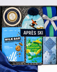 A curated gift box set labeled 'APRÈS SKI' containing a light blue knit beanie with a dark blue pom-pom, a blue and green striped knitted mini bag tied with a silver ribbon, a tin of Cheesecake & Honey spreads, a bag of Milk Bar pretzel chocolate chip cookies, a bag of sour elderflower fish, and a Ritual mountain blend chocolate bar, all arranged in gray packing material with blue satin fabric at the side.