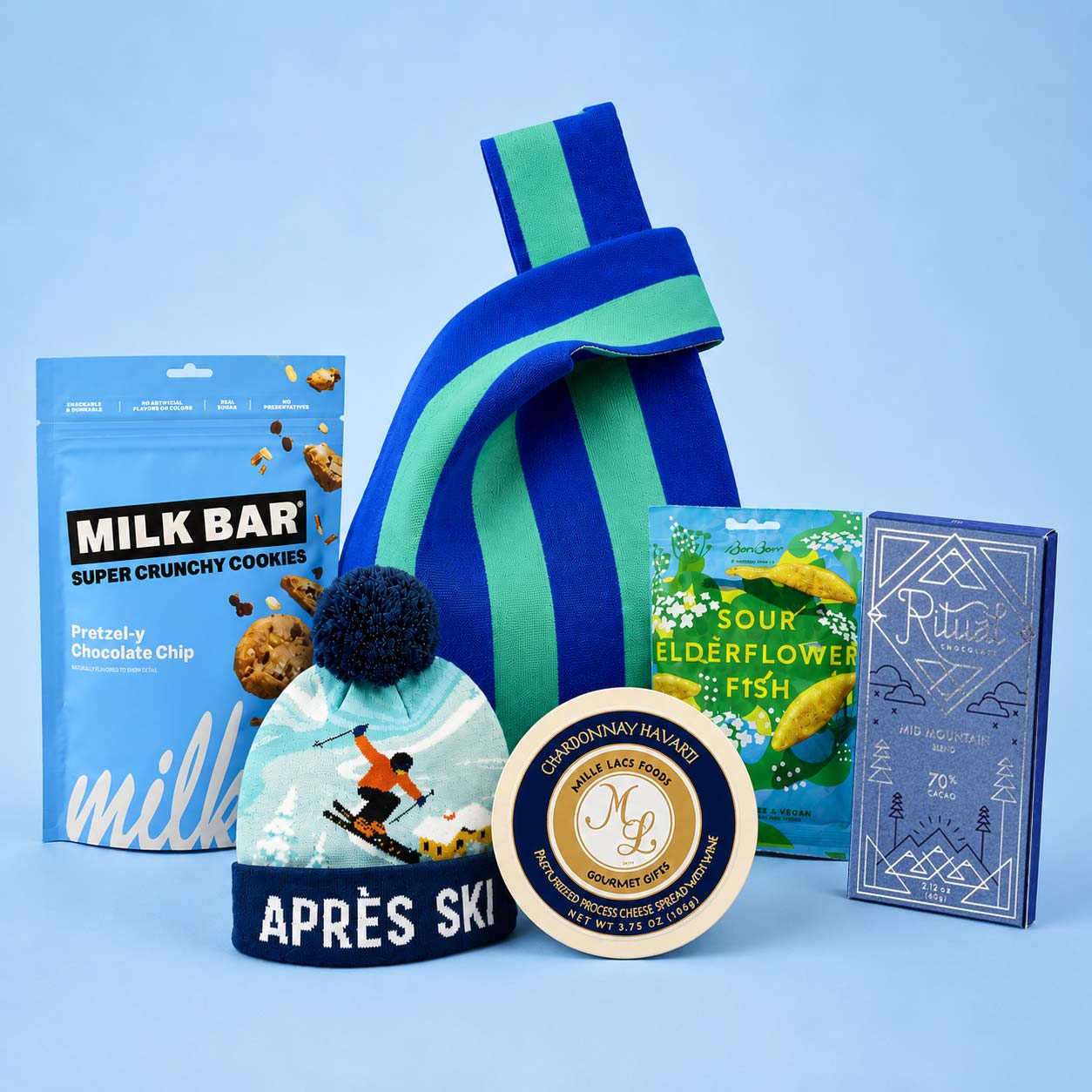 A curated gift set featuring a blue and green striped knit bag, a blue bag of Milk Bar super crunchy cookies, a round tin of chardonnay cheese, a box of Sour Elderflower &amp; Blue Fish, and a Ritual chocolate bar, all arranged on a light blue background.
