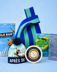 A curated gift set featuring a blue and green striped knit bag, a blue bag of Milk Bar super crunchy cookies, a round tin of chardonnay cheese, a box of Sour Elderflower & Blue Fish, and a Ritual chocolate bar, all arranged on a light blue background.