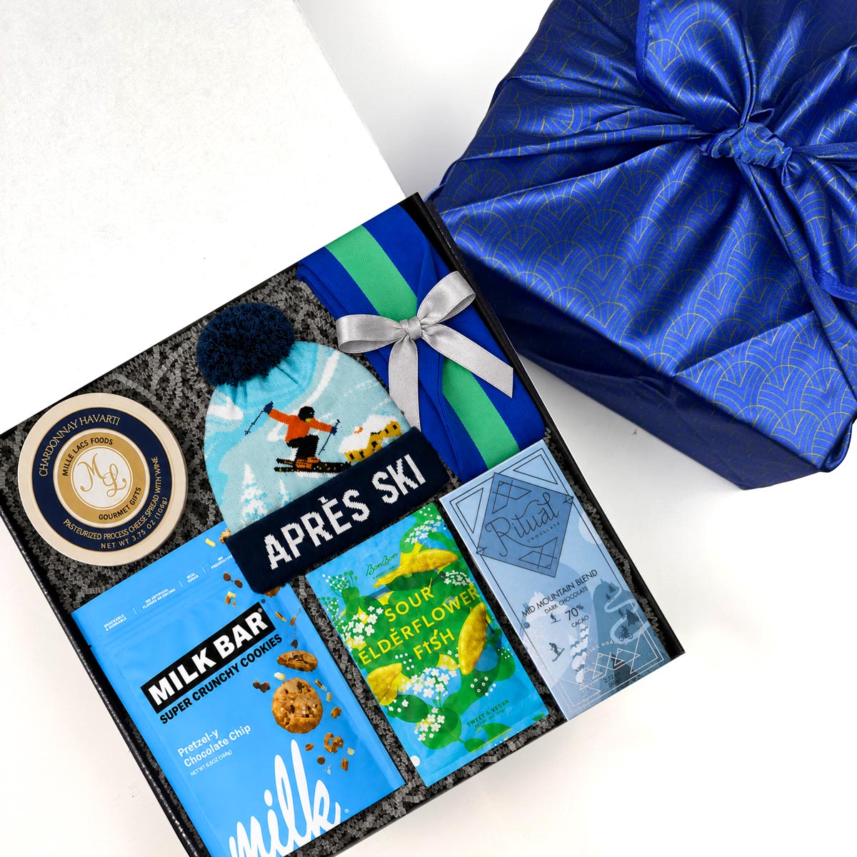 A curated gift box set on a white background, featuring a black gift box filled with winter-themed treats and accessories including a blue ski hat, chocolate bar, milk bar, and a blue green knitted bag.