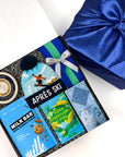 A curated gift box set on a white background, featuring a black gift box filled with winter-themed treats and accessories including a blue ski hat, chocolate bar, milk bar, and a blue green knitted bag.