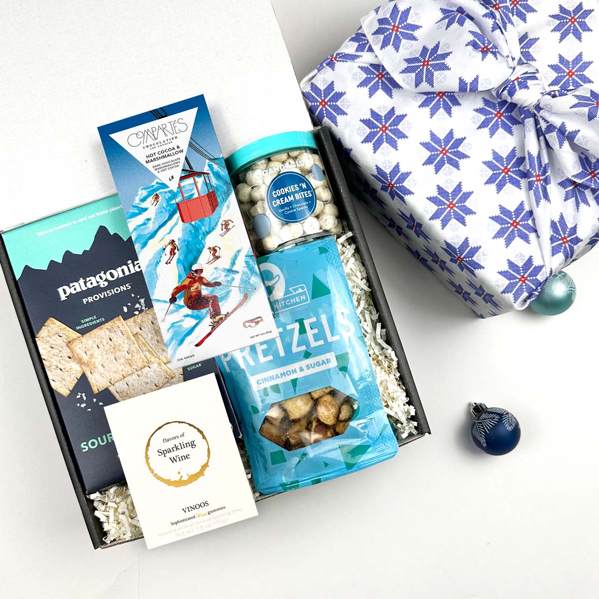 kadoo après ski holiday curated gift box in blue snowflake furoshiki wrap. Inside gifts hot cocoa marshmallow chocolate, patagonia rackers, vinoss wine gummies, pretzel, cookie cream bites and more