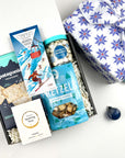 kadoo après ski holiday curated gift box in blue snowflake furoshiki wrap. Inside gifts hot cocoa marshmallow chocolate, patagonia rackers, vinoss wine gummies, pretzel, cookie cream bites and more