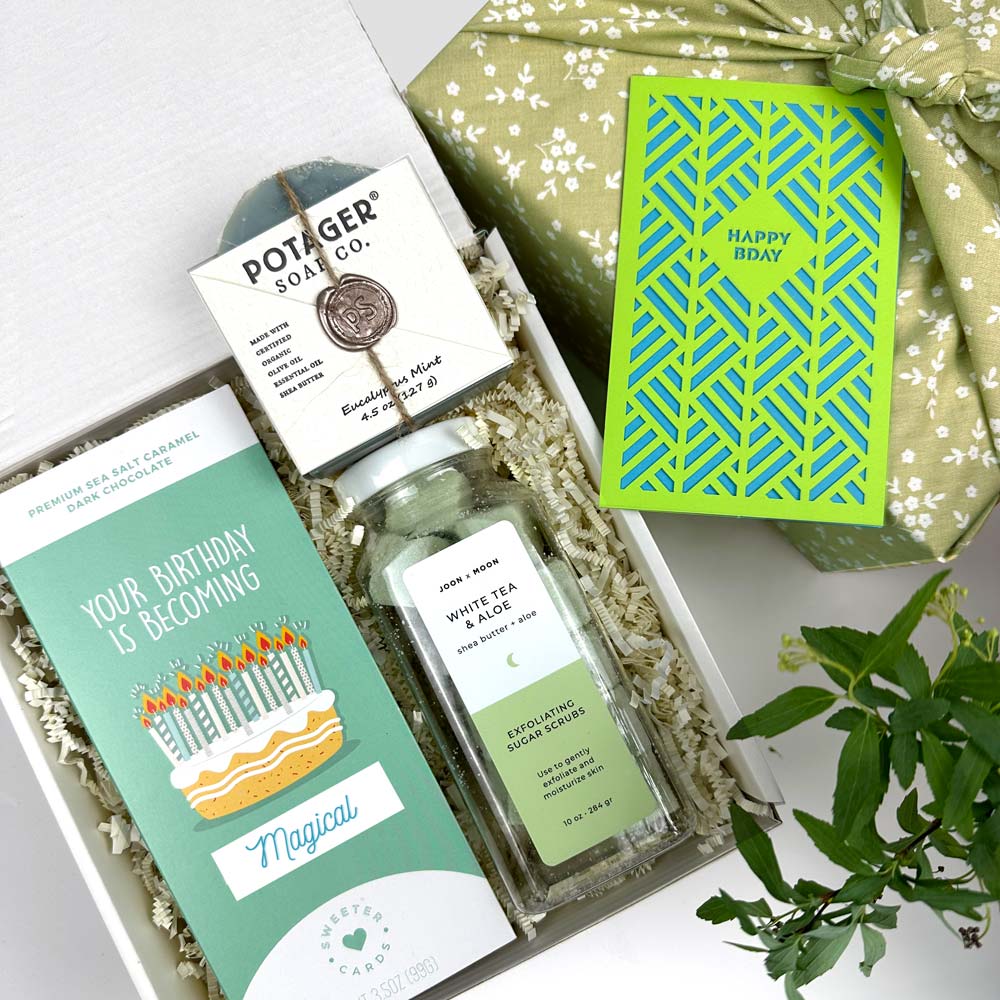 kadoo birthday celebration gift box. Inside gifts: birthday cake chocolate bar, soap eucalyptus mint, white tea aloe sugar scrub, happy birthday laser cut notecard in green and blue. Wrapped in green floral pattern gift box.