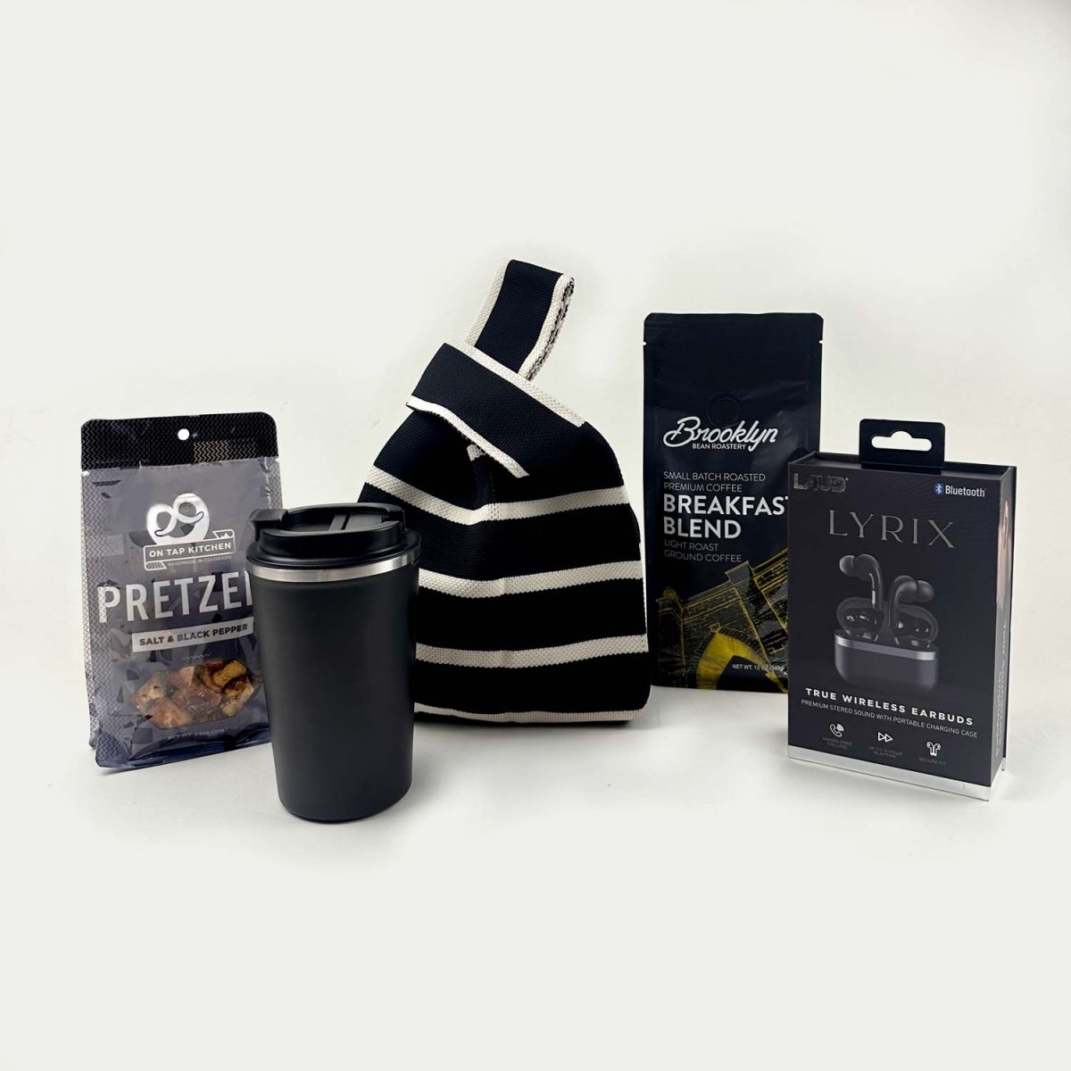 Black and white striped bag, black tumbler, pretzel packaging, coffee packaging, and wireless earbuds packaging on a white background.