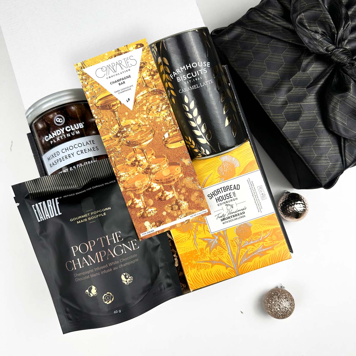 kadoo celebration holiday curated gift box with black furoshiki wrap. Inside gifts: pop the champagne popcorn, champagne chocolate bar, caramel latte biscuits, shortbread, chocolate raspberry cremes and more