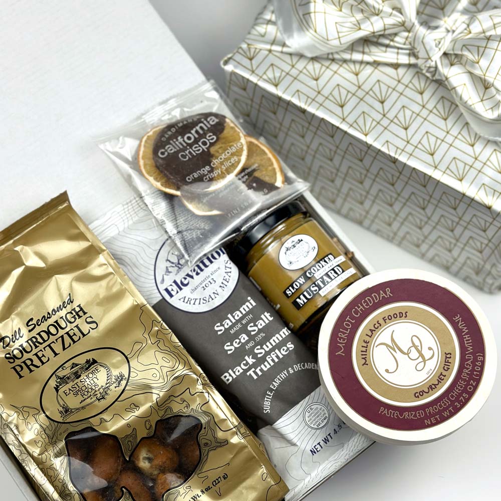 kadoo cheese and charcuterie gift box in gold white fabric wrap. Inside gift box: sourdough pretzels, salami, orange chocolate slice, mustard, cheese spread, and more. 