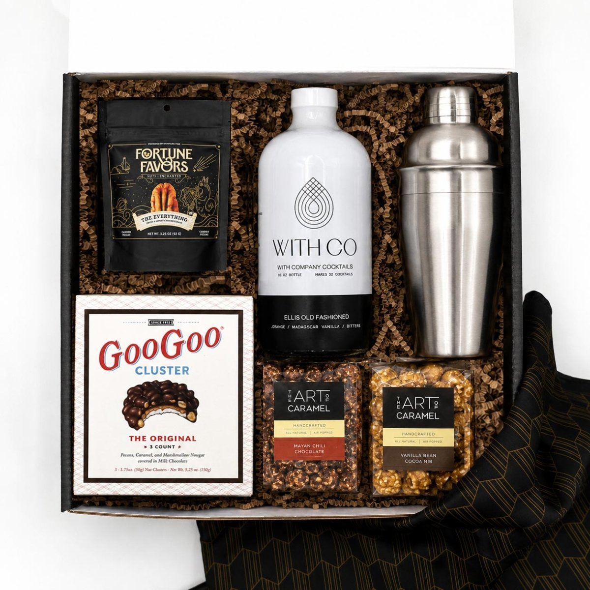 A curated gift box set containing a white bottle of With Co Old Fashioned Cocktails, a stainless steel shaker, a bag of Fortune Favors peanuts, a box of GooGoo Cluster chocolate, and two bags of Art of Caramel snacks, arranged in brown crinkle paper inside a black box.