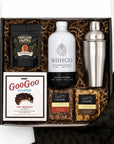 A curated gift box set containing a white bottle of With Co Old Fashioned Cocktails, a stainless steel shaker, a bag of Fortune Favors peanuts, a box of GooGoo Cluster chocolate, and two bags of Art of Caramel snacks, arranged in brown crinkle paper inside a black box.