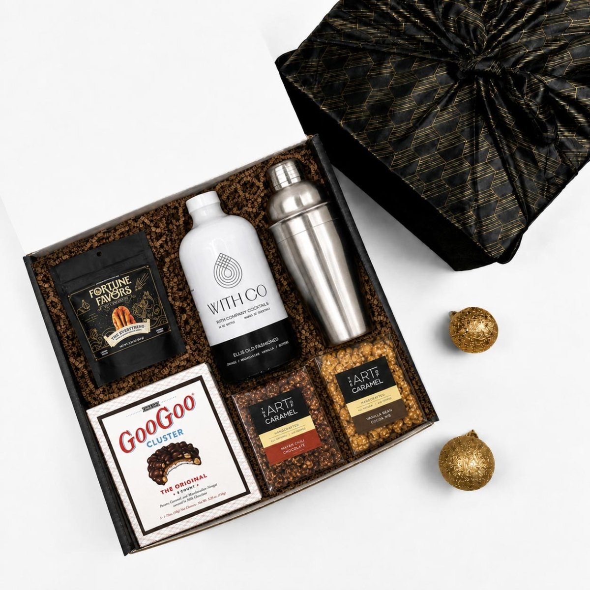 A curated gift box set on a white background, featuring a black and gold patterned gift box with its lid off, containing a white bottle labeled WITH CO, a stainless steel shaker, a bag of Fortune Favors Peanuts, a cookie box, and two bags of popocorns. Two gold decorative ornaments are placed beside the box.