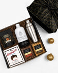 A curated gift box set on a white background, featuring a black and gold patterned gift box with its lid off, containing a white bottle labeled WITH CO, a stainless steel shaker, a bag of Fortune Favors Peanuts, a cookie box, and two bags of popocorns. Two gold decorative ornaments are placed beside the box.