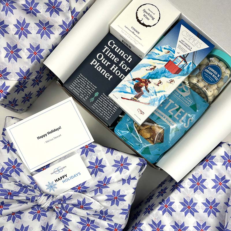 Gift box with holiday-themed items including a card, snacks, and a blanket with blue star pattern.