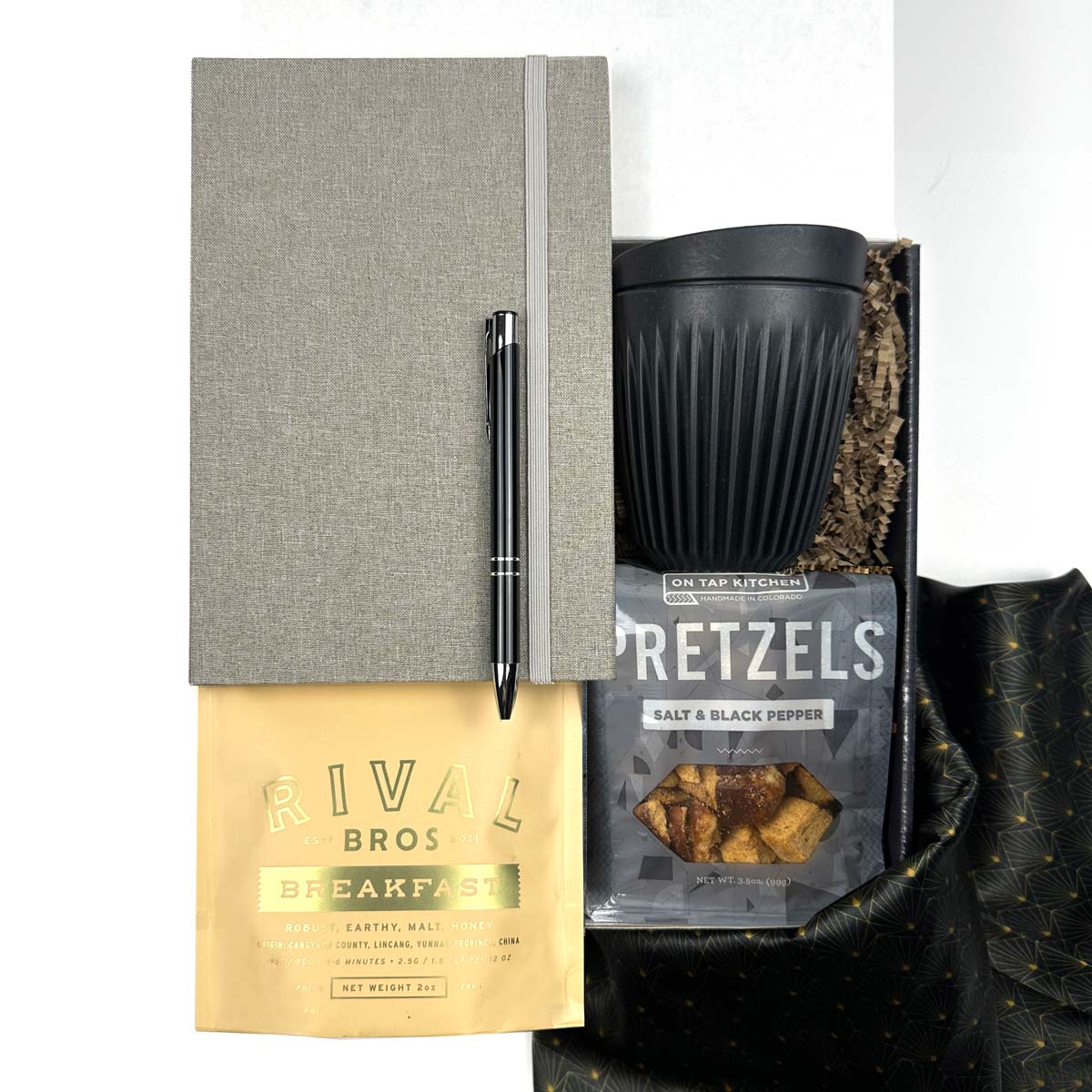 KADOO Desk essentials gift box with gray journal, black silver pen, breakfast tea, black mug, pretzels, in black gold fabric wrap and more.