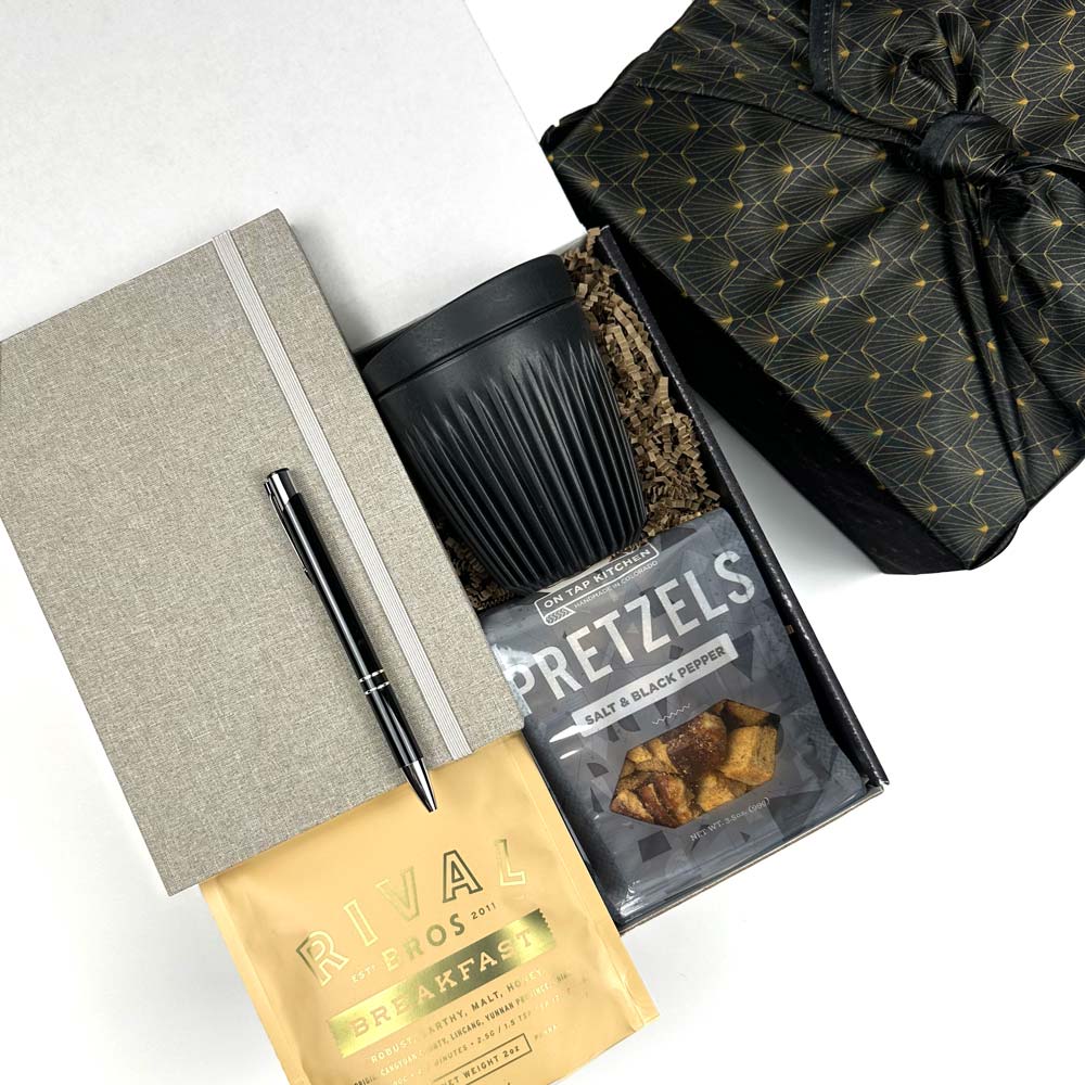 KADOO Desk essentials gift box with gray journal, black silver pen, breakfast tea, black mug, pretzels. Wrapped in black gold furoshiki style fabric wrap.