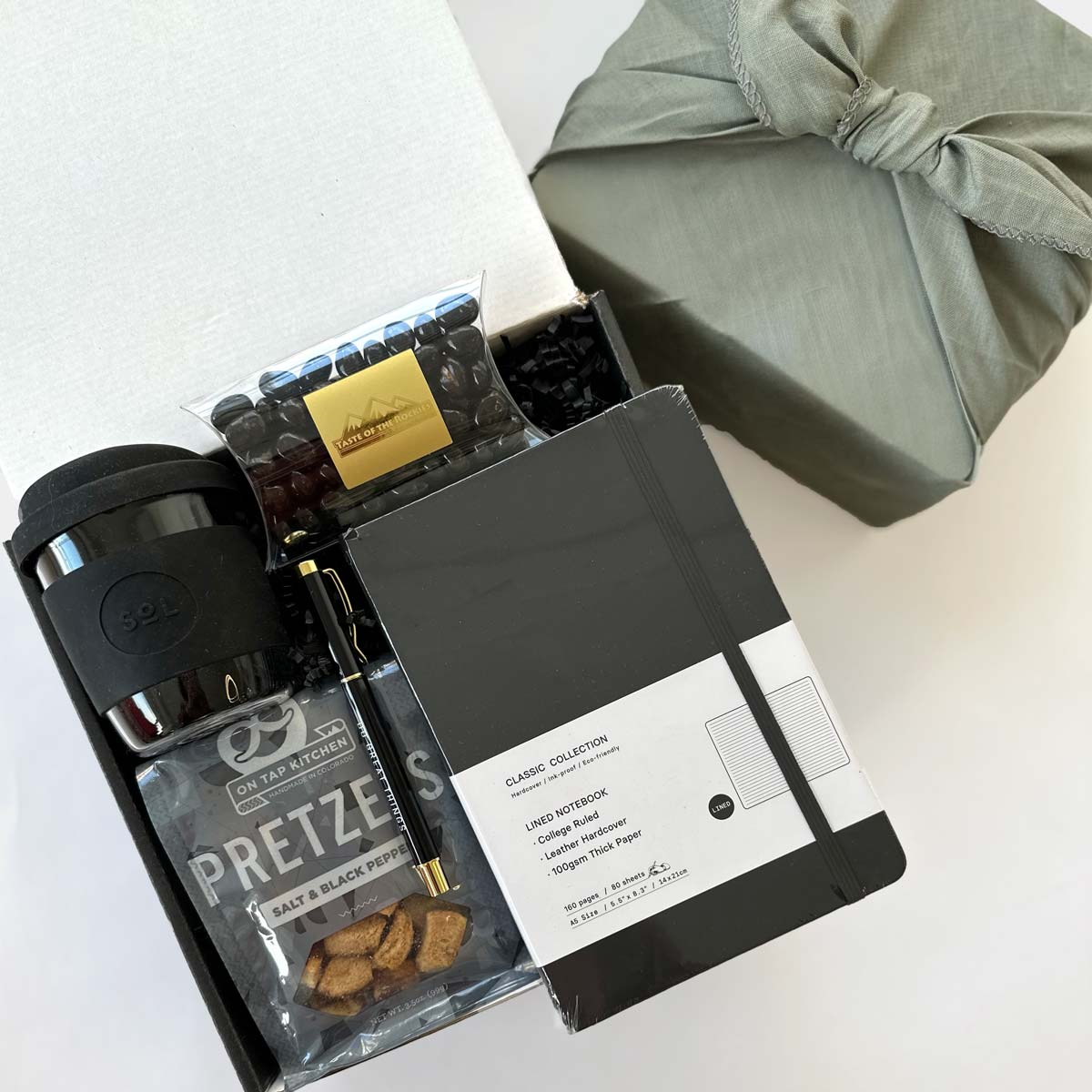kadoo do great things gift box with black sol cup, pretzel, black notebook, pen, espresso beans candy and more. wrapped in gray fabric furoshiki wrap.