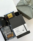 kadoo do great things gift box with black sol cup, pretzel, black notebook, pen, espresso beans candy and more. wrapped in gray fabric furoshiki wrap.