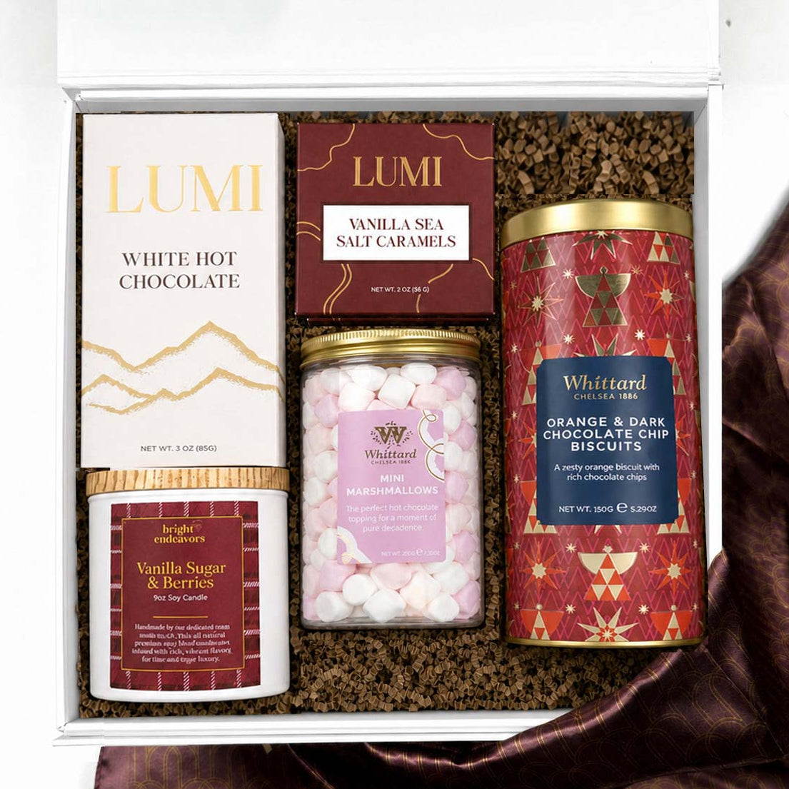 An open gift box containing curated treats: a white hot chocolate , vanilla sea salt caramels, vanilla sugar berries candle,  jar of mini marshmallow, and an orange and dark chocolate chip cookie tin, arranged in brown crinkle paper on a white background with a burgundy fabric accent.