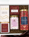 An open gift box containing curated treats: a white hot chocolate , vanilla sea salt caramels, vanilla sugar berries candle,  jar of mini marshmallow, and an orange and dark chocolate chip cookie tin, arranged in brown crinkle paper on a white background with a burgundy fabric accent.