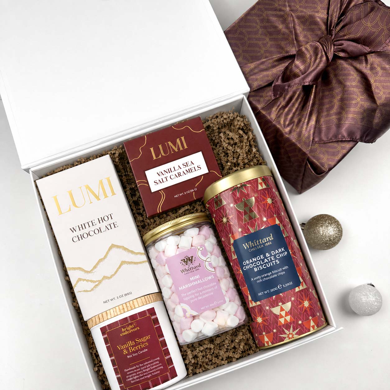 A curated gift box set containing a box of Lumi White Hot Chocolate, a jar of Whittard marshmallows, and a tin of Whittard Orange Grands & Dark Chocolates, a candle, box of lumi caramels, arranged in a white box with brown crinkle paper filler, alongside a wrapped gift and holiday ornaments.